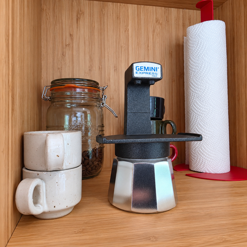 A Black Gemini Express moka pot coffee maker sits on a wooden shelf next to stacked ceramic mugs, a glass jar of coffee beans, and a roll of paper towels on a red holder.