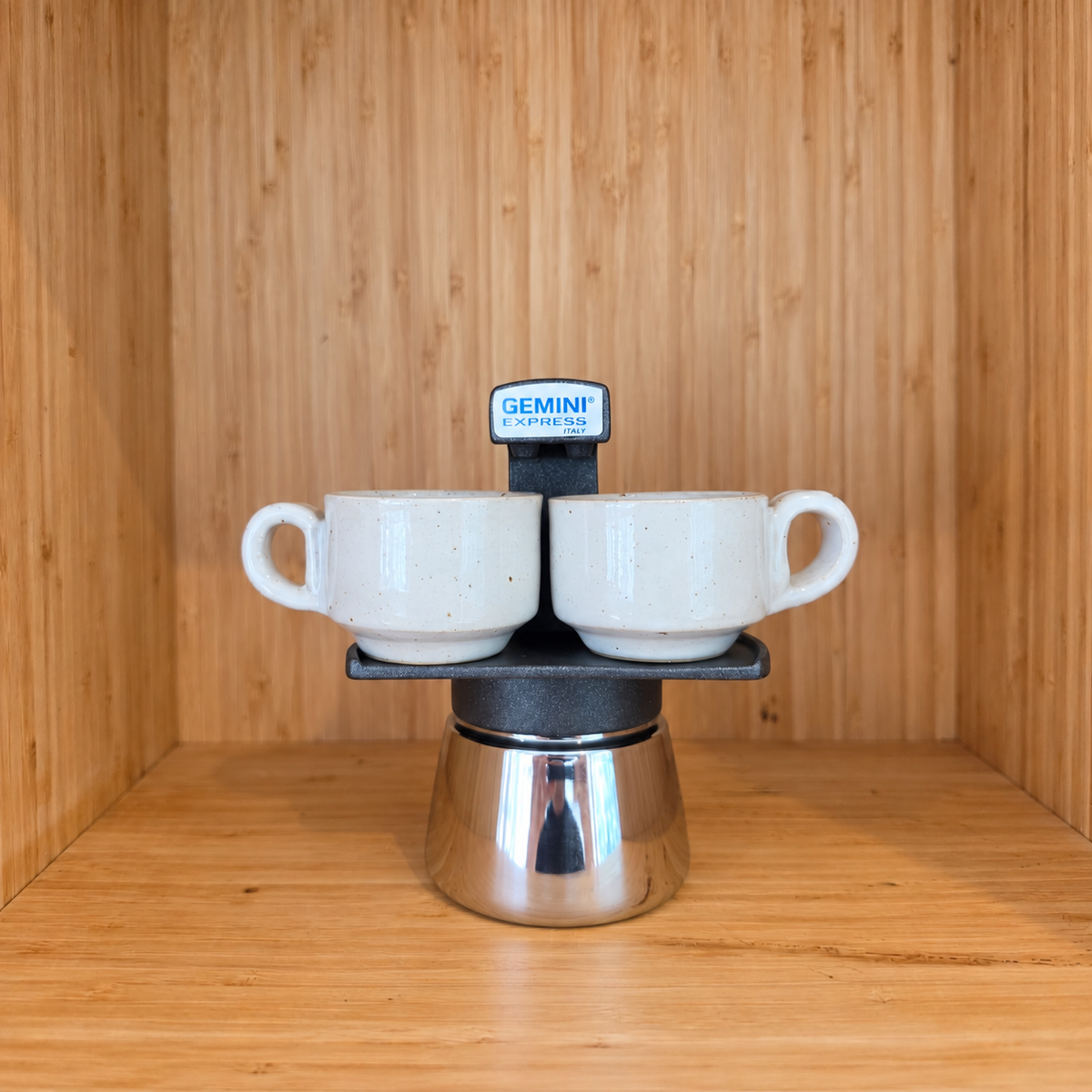 A Black Gemini Express moka pot centered with 2 mugs against a wooden background.
