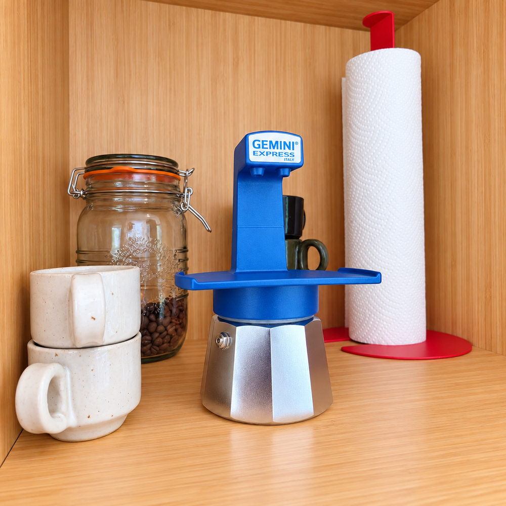 A Gemini Espresso moka pot coffee maker displayed on a wooden shelf next to stacked ceramic mugs, a glass jar of coffee beans, and a paper towel holder.