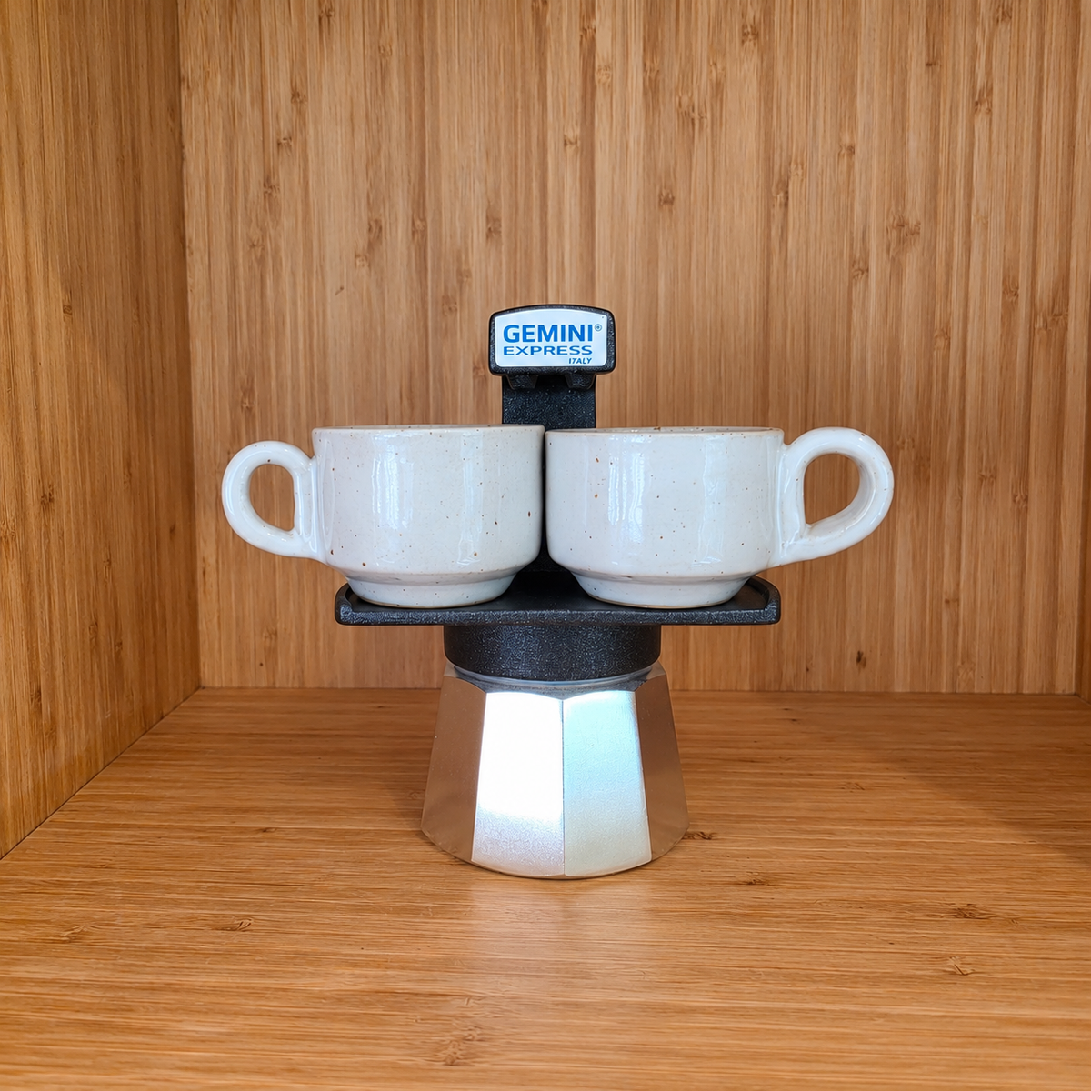 A Black Gemini Express moka pot centered with 2 mugs against a wooden background.