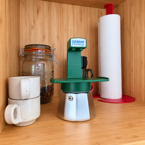 A green Gemini Express moka pot sits on a wooden shelf next to stacked cream mugs, a glass jar of coffee beans, and a paper towel holder with a roll of paper towels.