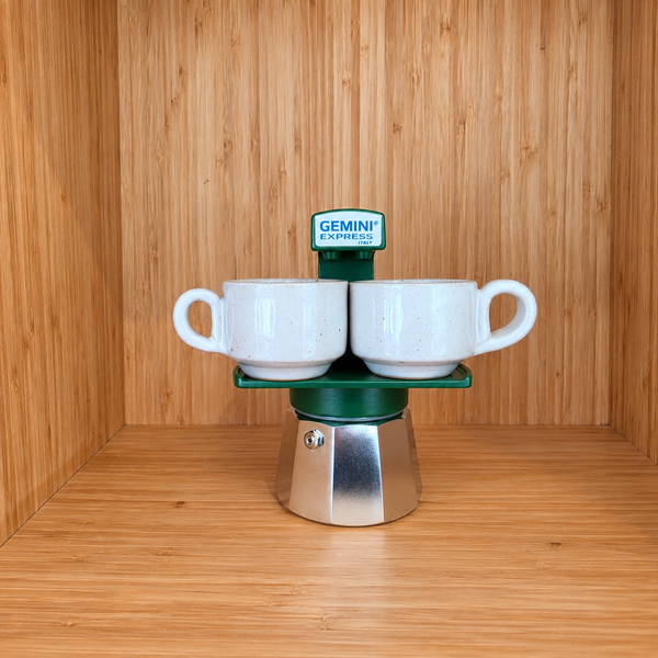 A green Gemini Express moka pot centered with 2 mugs on a wooden surface against a wooden wall background.