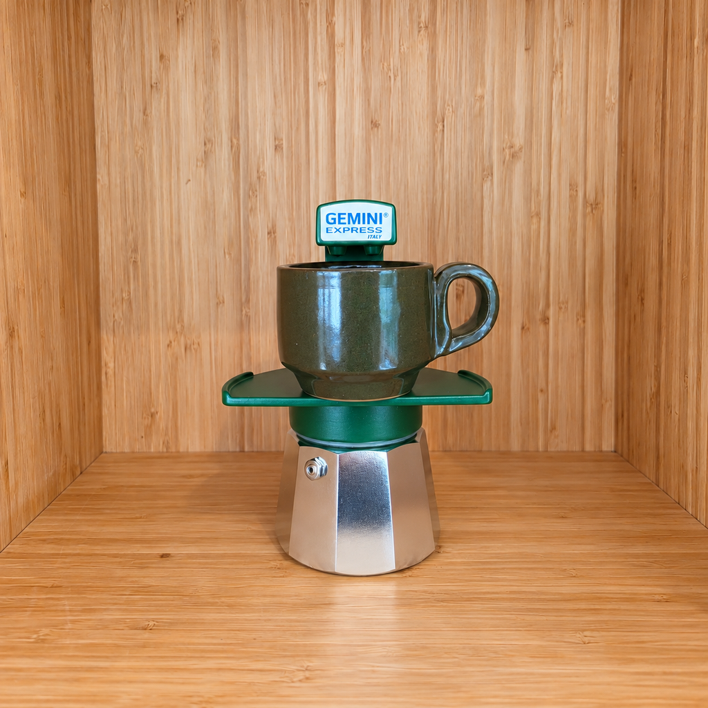 A green Gemini Express moka pot centered with 1 mug on a wooden surface against a wooden wall background.