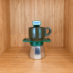 A green Gemini Express moka pot centered with 1 mug on a wooden surface against a wooden wall background.
