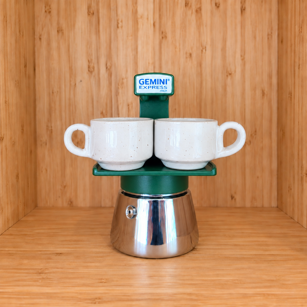 A Green Gemini Express espresso moka pot centered with 2 mugs on a wooden surface against a wooden wall background.