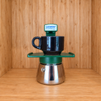 A Green Gemini Express espresso moka pot centered with 1 mug on a wooden surface against a wooden wall background.