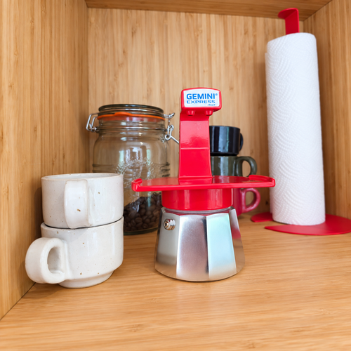 A Red Gemini Express Moka pot sits on a wooden shelf next to stacked white ceramic mugs, a glass jar of coffee beans, and a paper towel holder.
