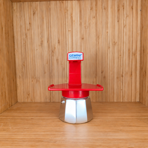 A red Gemini Espresso moka pot centered on a wooden shelf against a wooden wall.