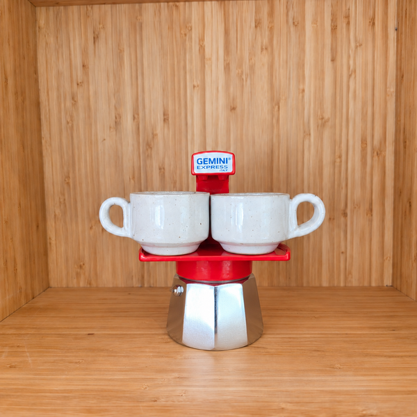 A red Gemini Espresso moka pot centered with 2 mugs on a wooden shelf against a wooden wall.