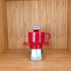 A red Gemini Espresso moka pot centered with a red mug on a wooden shelf against a wooden wall.