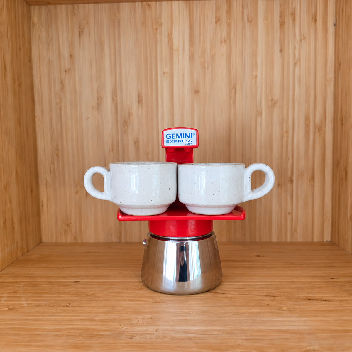 A red Gemini Espresso moka pot centered with 2 mugs on a wooden shelf against a wooden wall.