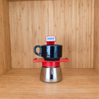 A red Gemini Espresso moka pot centered with a blue mug on a wooden shelf against a wooden wall.