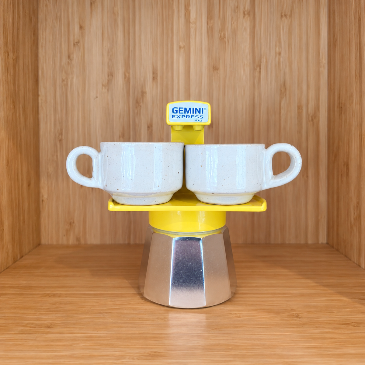 A Yellow Gemini Express espresso moka pot centered with 2 mugs on a wooden surface against a wooden wall background.