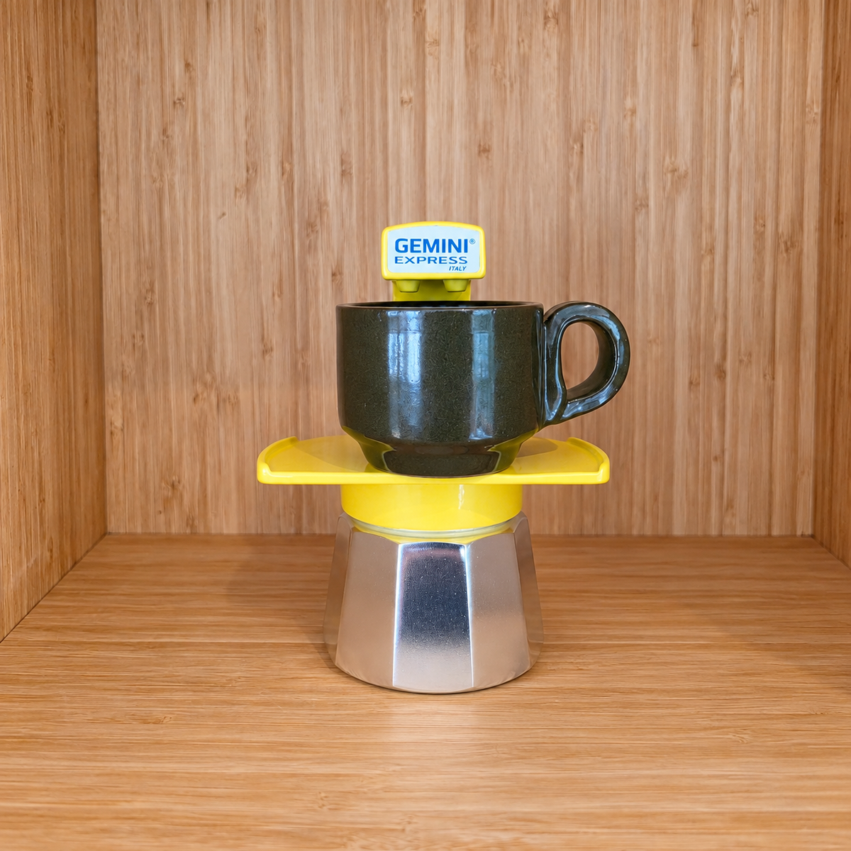 A Yellow Gemini Express espresso moka pot centered with 1 mug on a wooden surface against a wooden wall background.
