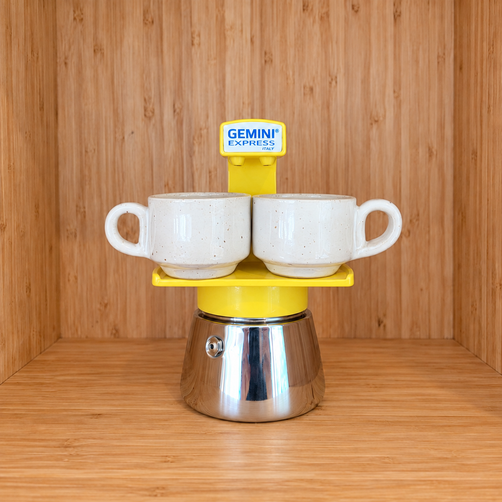 A yellow Gemini Express espresso moka pot centered with 2 mugs on a wooden surface against a wooden wall background.