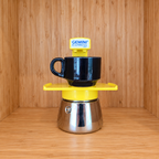 A yellow Gemini Express espresso moka pot centered with one mug on a wooden surface against a wooden wall background.