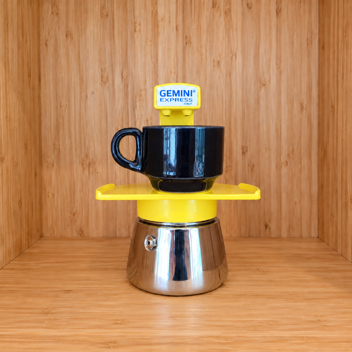 A yellow Gemini Express espresso moka pot centered with one mug on a wooden surface against a wooden wall background.
