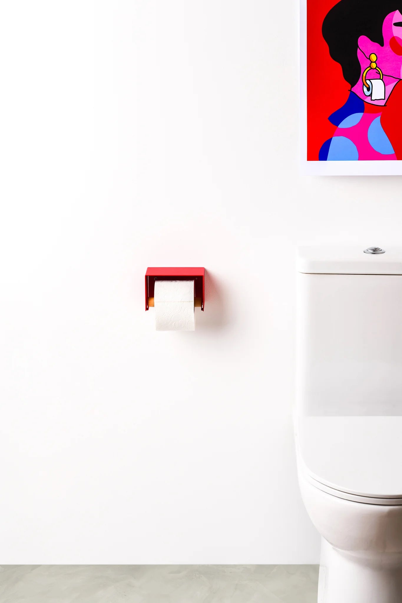 A modern red wall-mounted toilet paper holder with a white roll of toilet paper, installed on a white bathroom wall next to a white toilet, with a colorful pop-art print hanging on the wall.