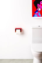 A modern red wall-mounted toilet paper holder with a white roll of toilet paper, installed on a white bathroom wall next to a white toilet, with a colorful pop-art print hanging on the wall.